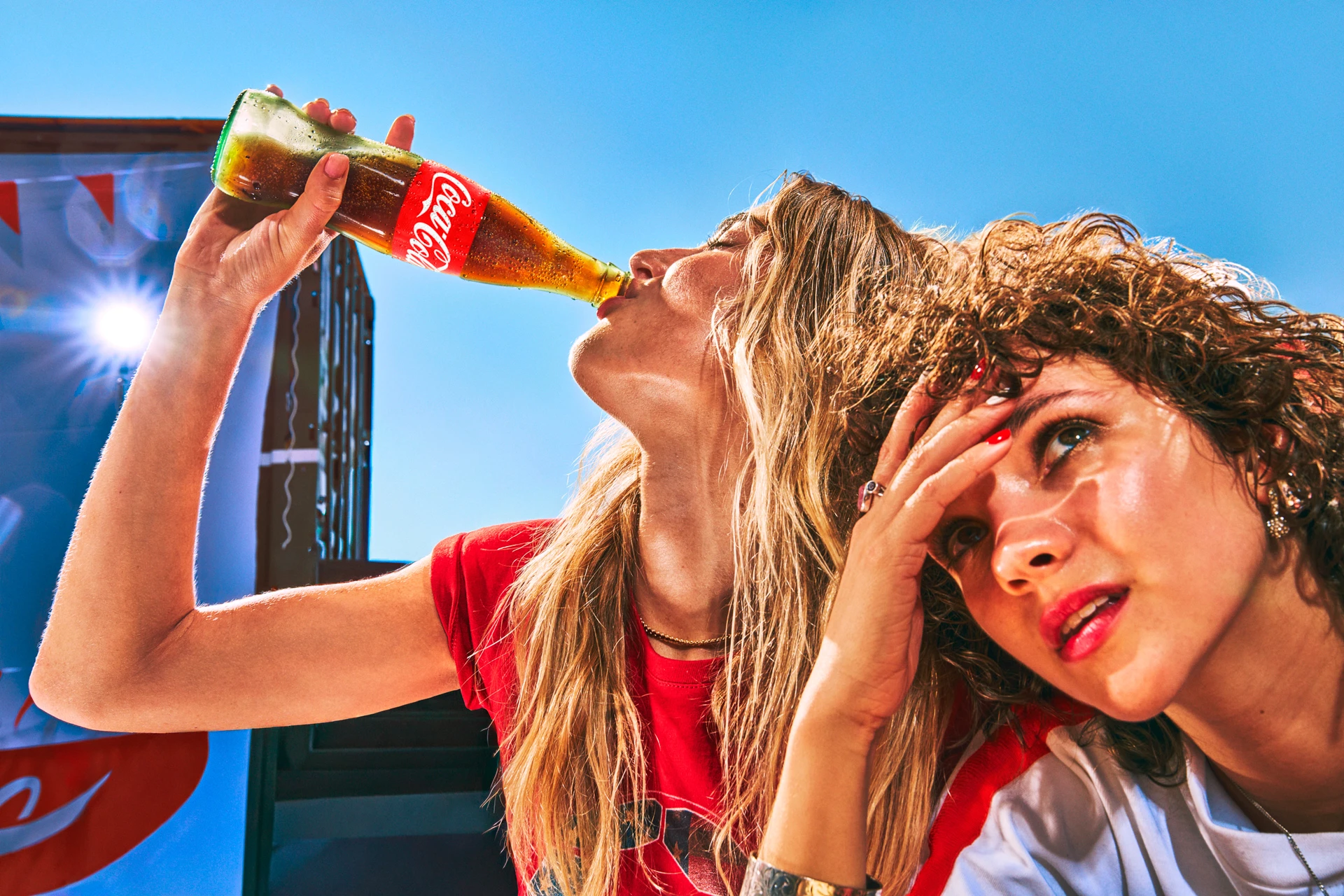 Zwei junge Frauen im Sommer im Freien, wobei eine von ihnen eine Coca-Cola Flasche ansetzt.